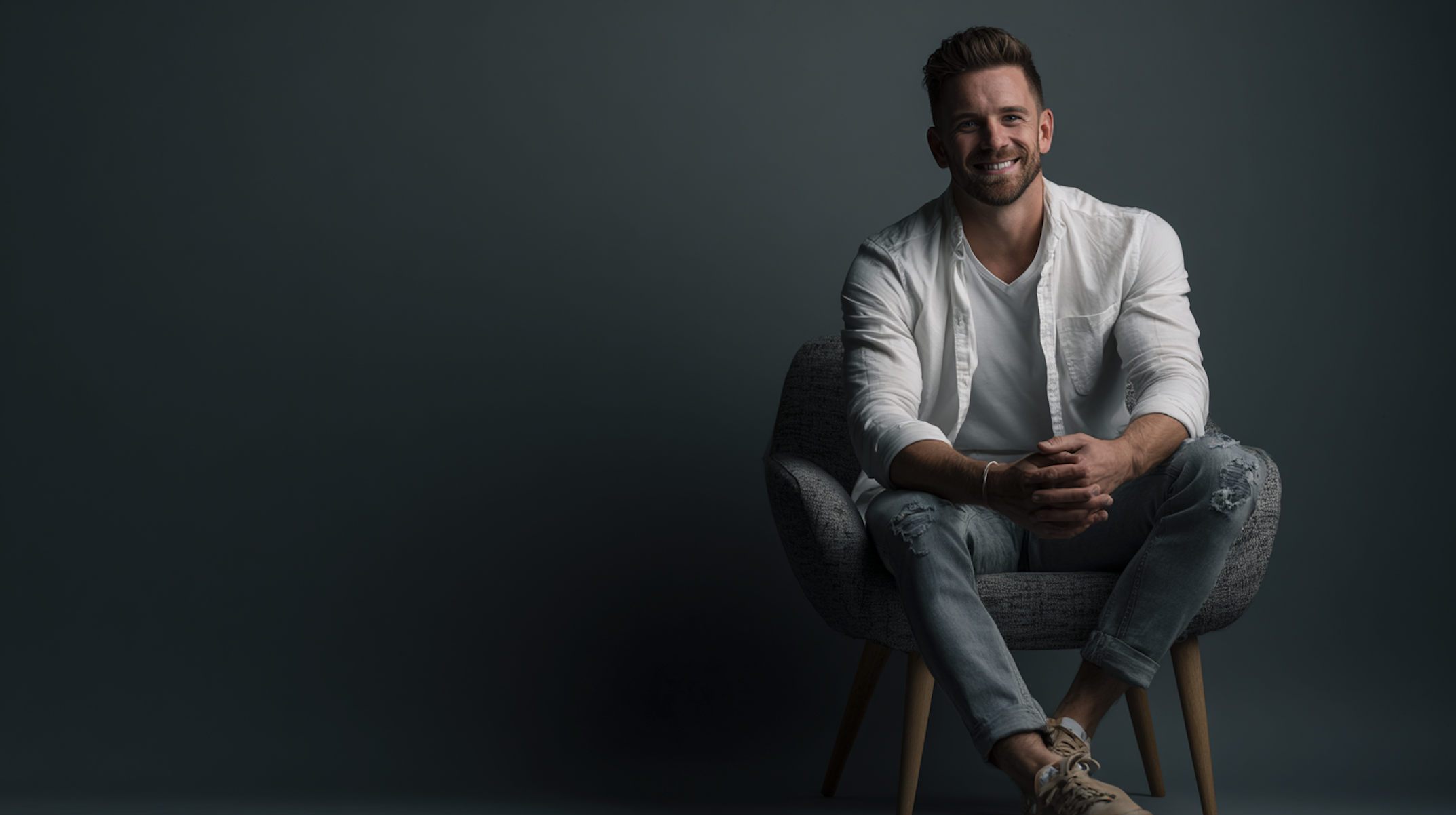 male wearing a white shirt and blue jeans sitting in front of a gray background